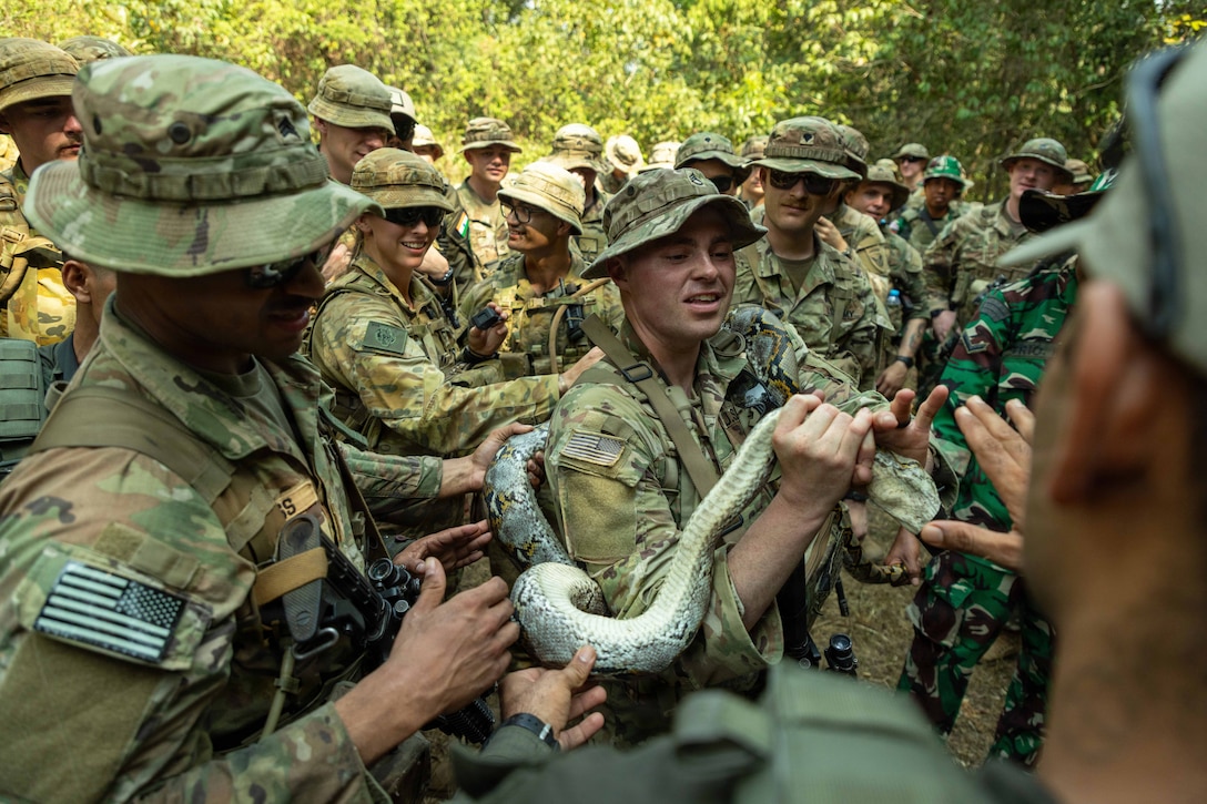 Dozens of soldiers in uniform in a wooded area gather around a fellow soldier holding a snake during the day.