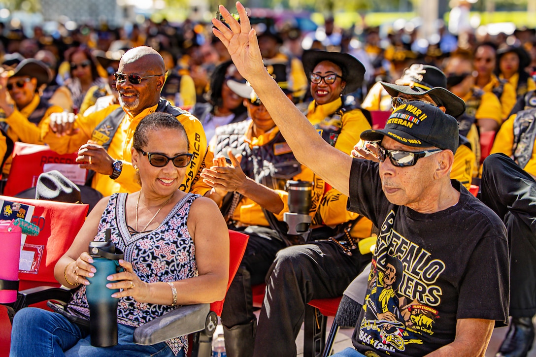 A 104-year-old veteran sitting to the right of a crowd waves as the audience wearing black and yellow smiles and applauds to the left during the day.
