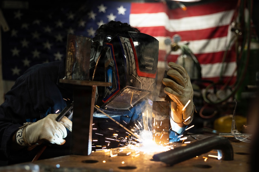 Sparks fly as a sailor wearing protective gear welds metal in a dimly lit room with an American flag hanging in the background.