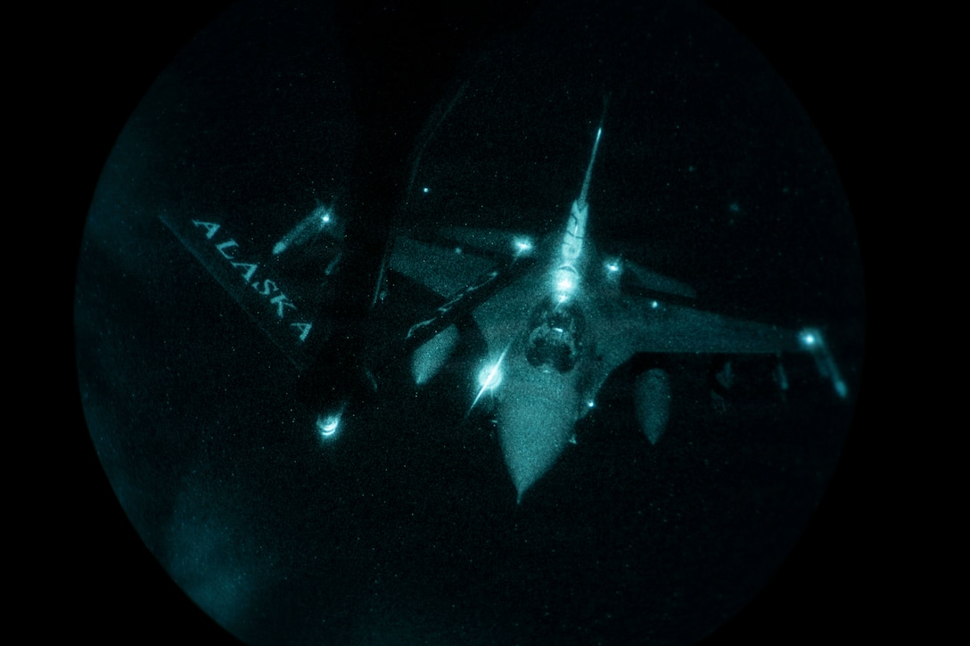 An airborne aircraft approaches the rear of another aircraft in the dark illuminated by its own lights.