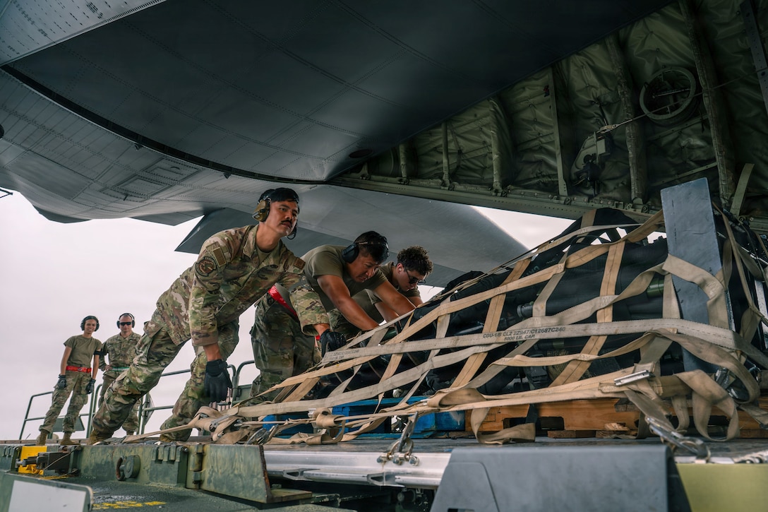 Three service members wearing camouflage uniforms push a pallet of equipment onto an aircraft while two others look on during daytime.