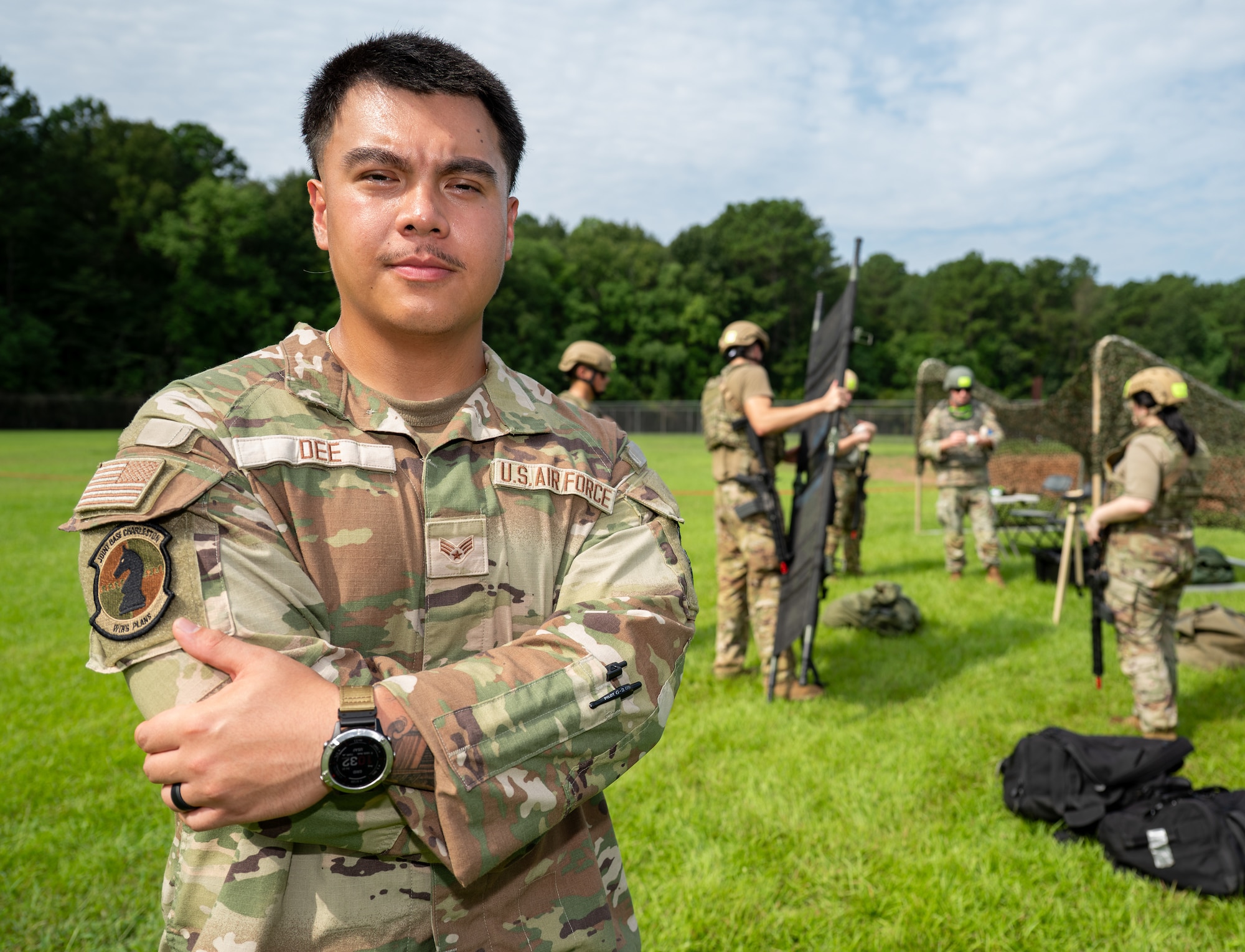 A man poses with his arms folded with Airmen in heavy gear in the background.