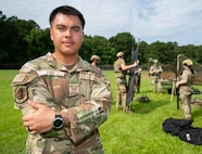 A man poses with his arms folded with Airmen in heavy gear in the background.