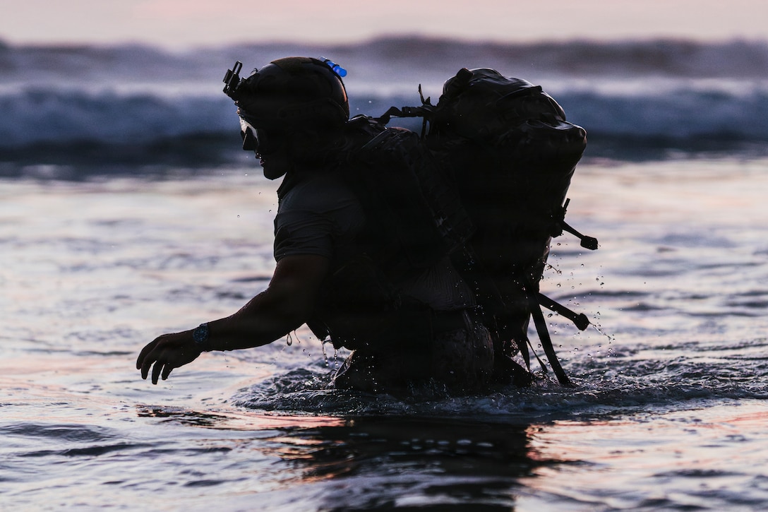A sailor wearing a helmet and backpack is silhouetted while wading through the surf in dim light conditions.