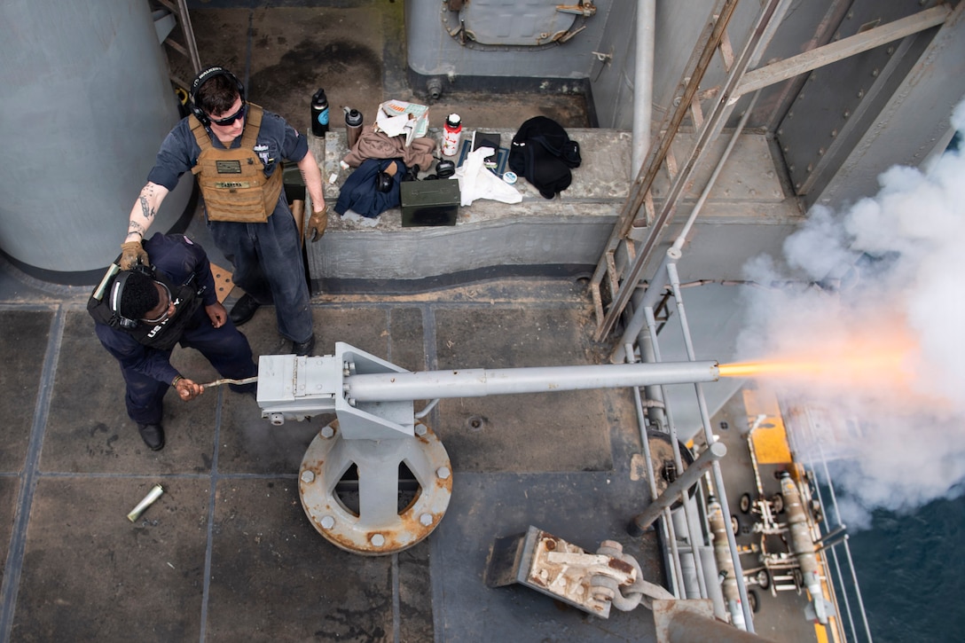 A sailor fires a cannon on a ship while another sailor stands to the side during daytime.