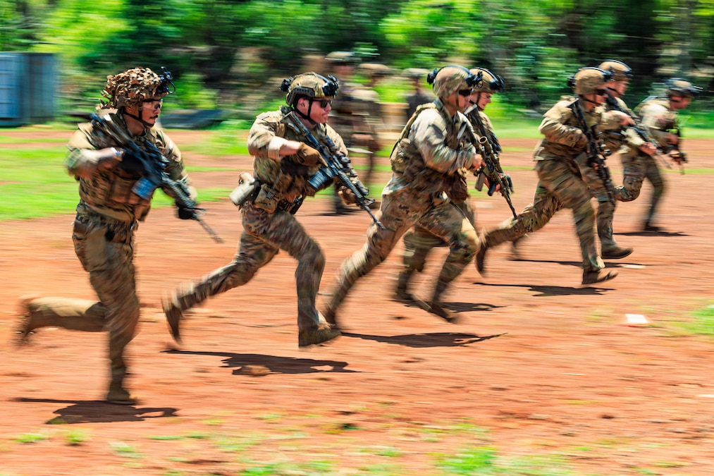 Seven soldiers wearing camouflage uniforms, helmets, and load-bearing equipment run while carrying their rifles on a bright day.