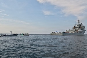 A large ship deploys a refueling rig using a cable toward an unmanned vessel sailing in water.