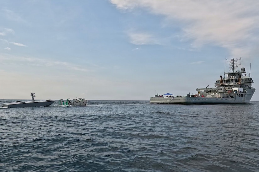 A large ship deploys a refueling rig using a cable toward an unmanned vessel sailing in water.