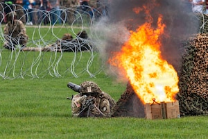 A person wearing combat gear lies in the grass holding a weapon while a wooden box next to him is on fire. There are people in similar attire in the background.