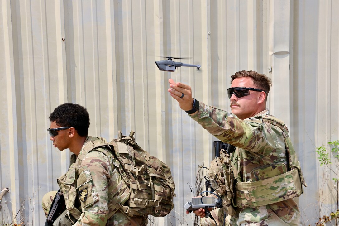 A small drone hovers above the outstretched arm of a soldier standing by a structure outdoors by another soldier.