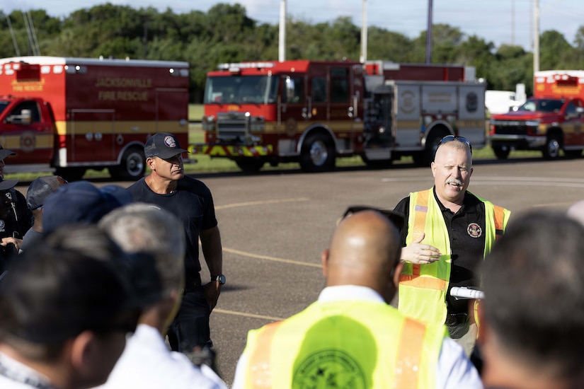 A man in business casual attire and a reflective vest talks to a dozen people in similar attire standing in a parking lot outside; there are three emergency vehicles parked in the background.