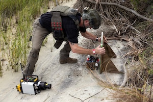 A man in emergency response gear puts an X-ray machine next to a simulated explosive sitting in the sand.
