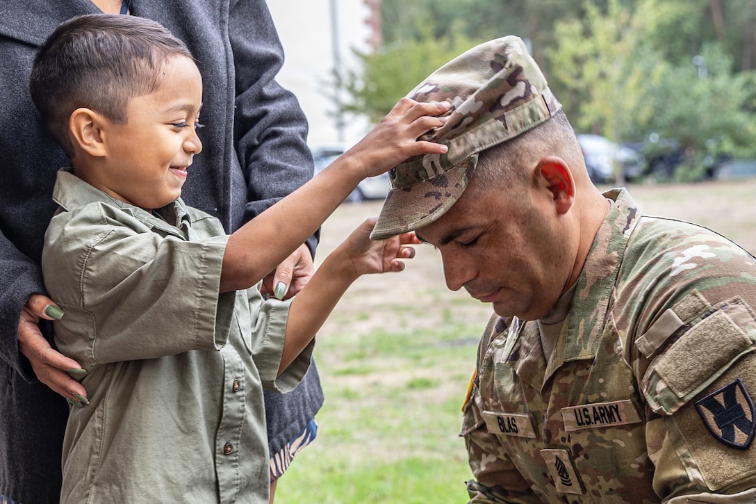 A smiling military child laughs while placing a hat on a kneeling soldier’s head.