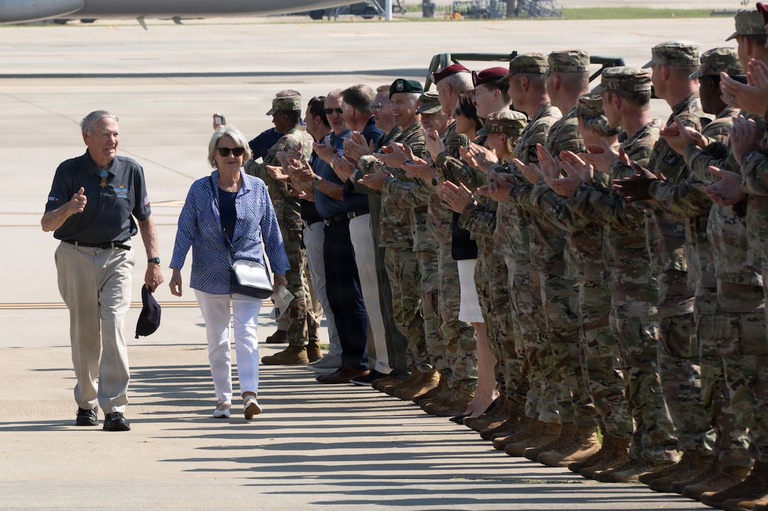 A veteran and their spouse walk up a ramp while soldiers standing in formation smile and applaud.