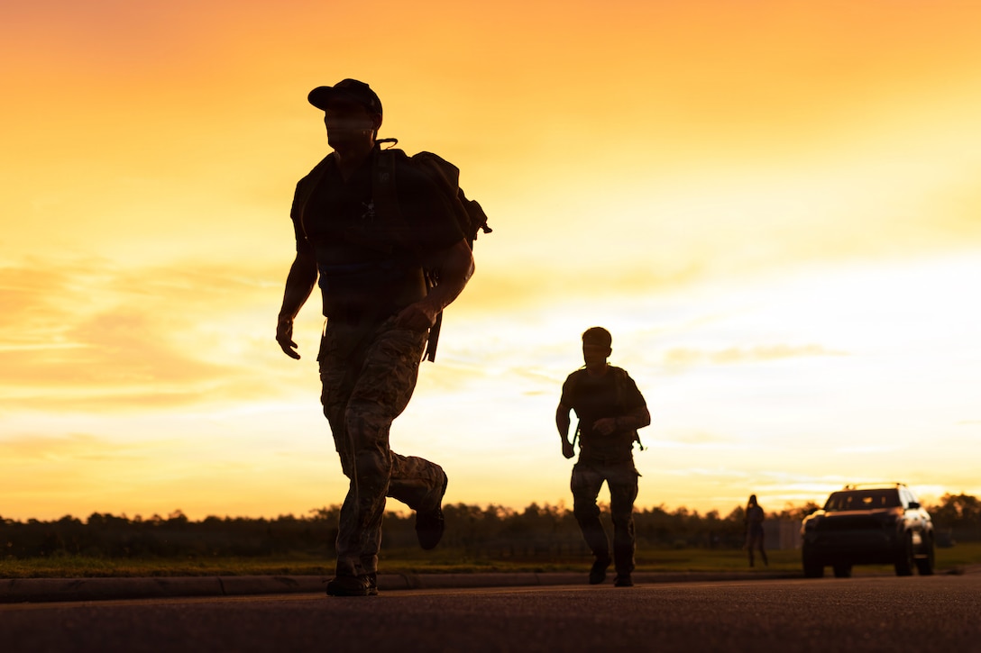 Two soldiers in uniform carrying backpacks are shown in silhouette while running as a golden sun fills the background.