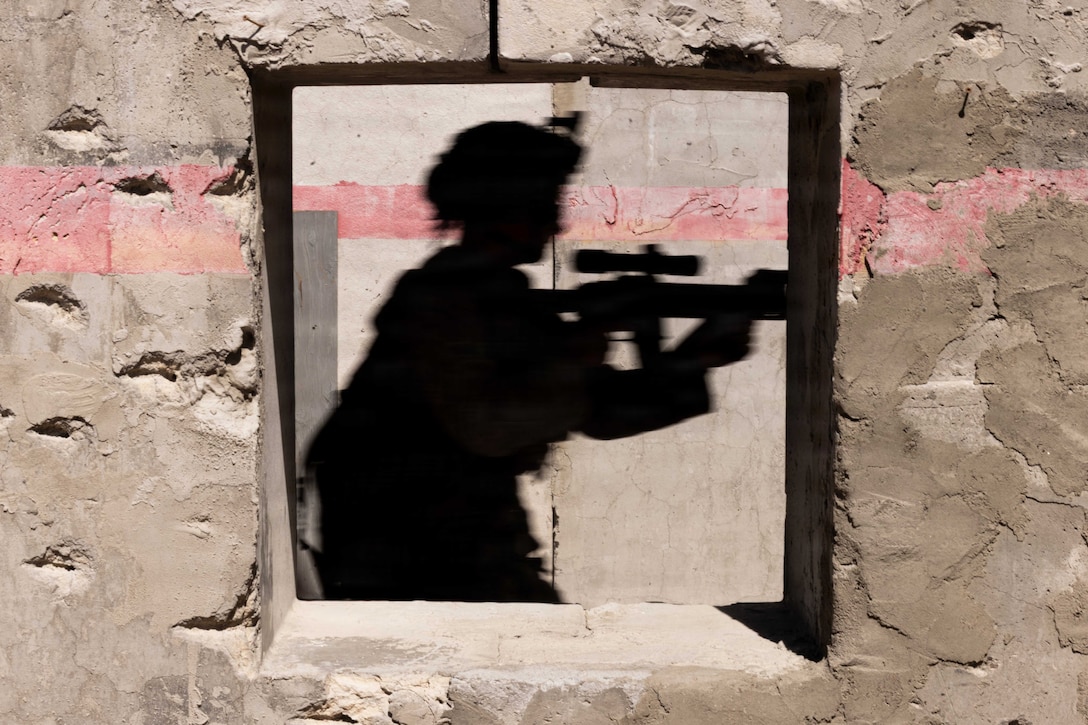 A shadow of a Marine in tactical gear aiming a weapon is seen on a window of a concrete wall.