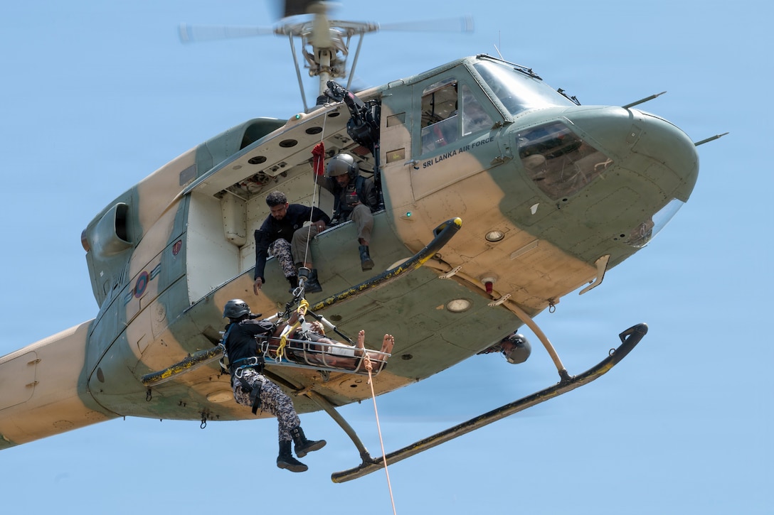 A simulated casualty on a rescue litter and a service member wearing camouflage pants and a black jacket are being pulled up into a hovering military helicopter by two service members on a bright day.