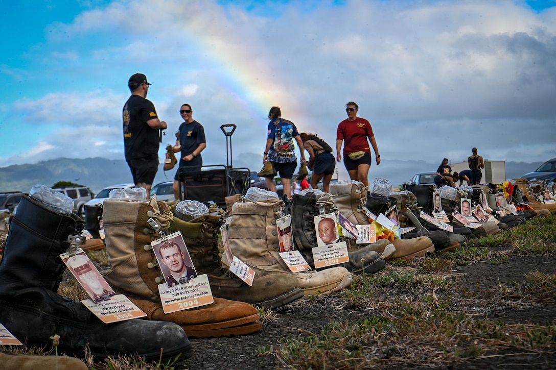 Ten people wearing casual clothing place combat boots with tags on them in lines outdoors under a rainbow on a partially cloudy day.