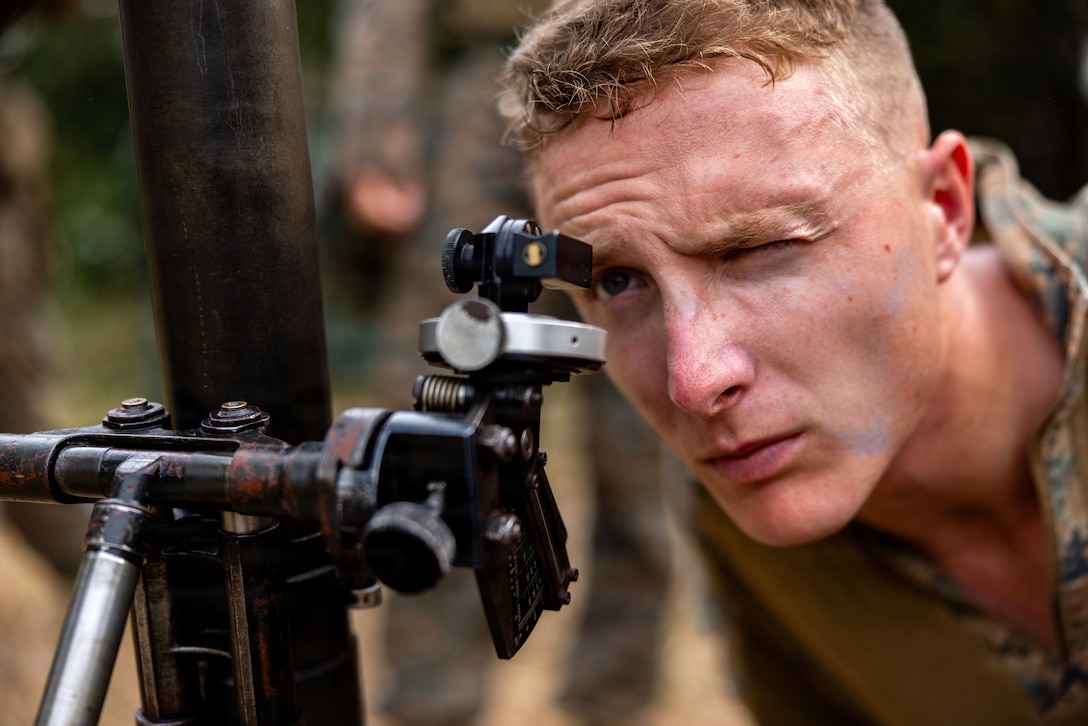 A person wearing a camouflage uniform closes one eye as he looks through a sight on a mortar.