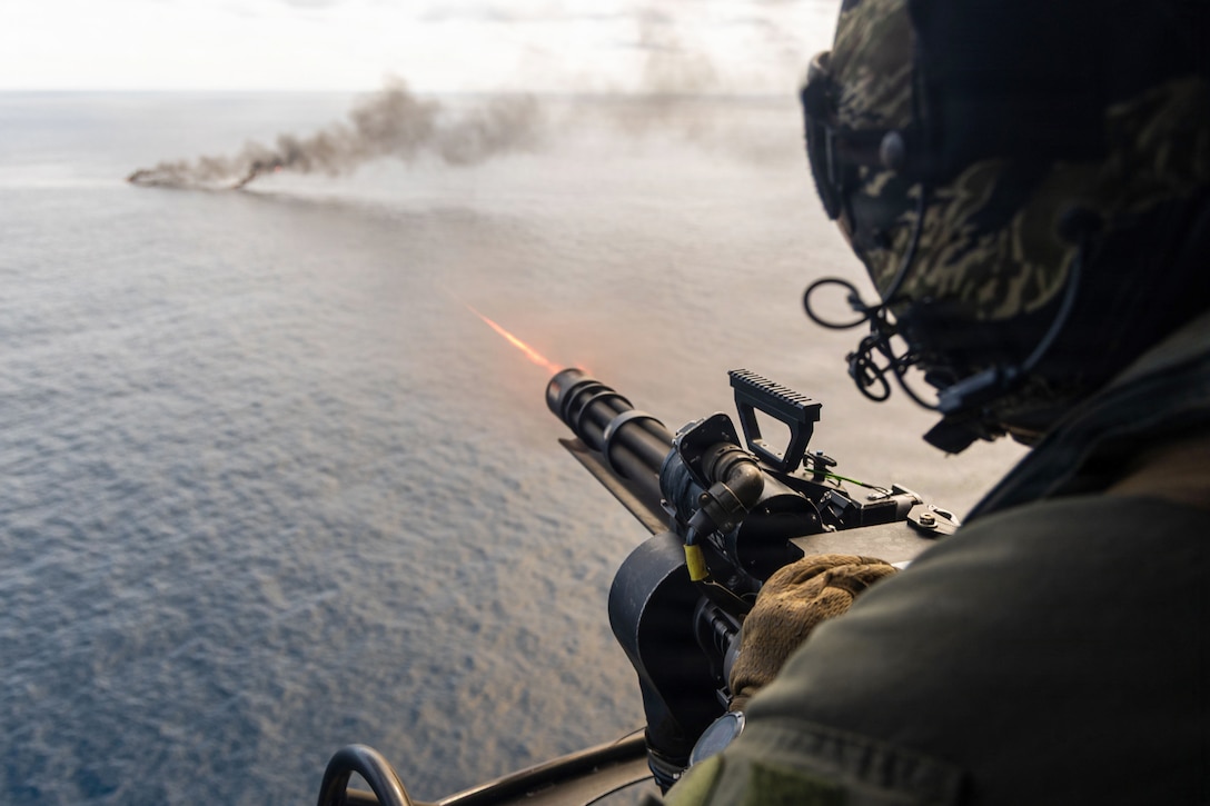 A person in a flying helicopter wearing a flight suit and a helmet fires a machine gun toward an object billowing smoke on a body of water.