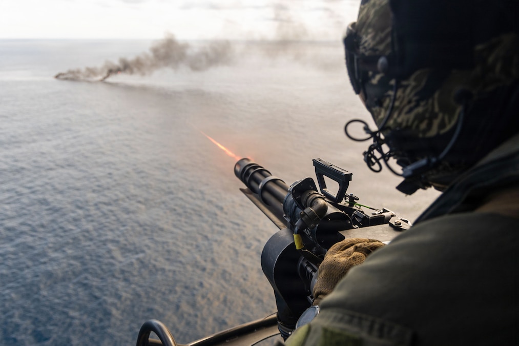 A person in a flying helicopter wearing a flight suit and a helmet fires a machine gun toward an object billowing smoke on a body of water.