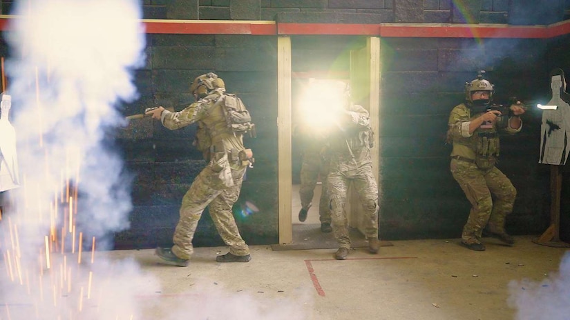 People in camouflage military uniforms and tactical gear walk through a door while having weapons drawn as a cloud of smoke and sparks cover the room.