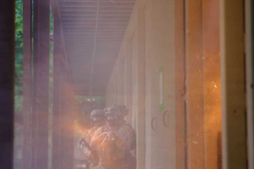 Three people in camouflage military uniforms and tactical gear walk in a hallway filled with smoke.