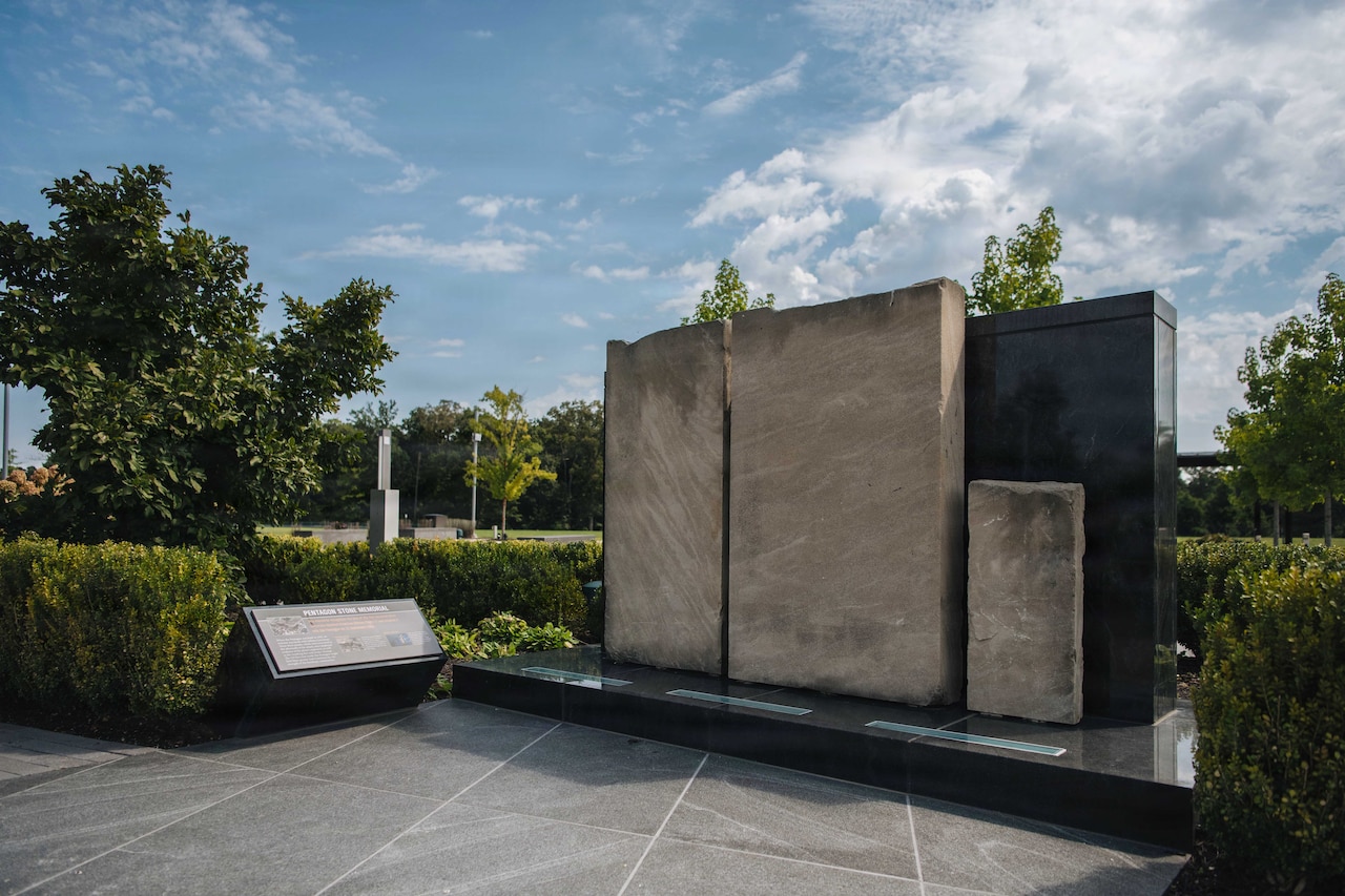 Three large slabs of stone are in a plaza area outside, with a slab with words on it next to them.