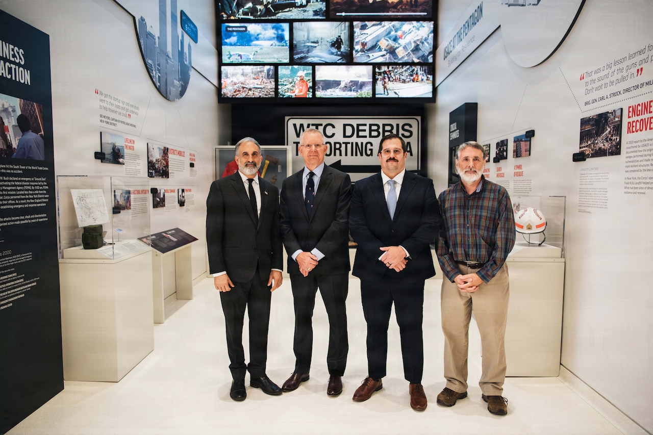 Three people in suits and one in business-casual attire pose for a photo in fron of 9/11 artifacts.