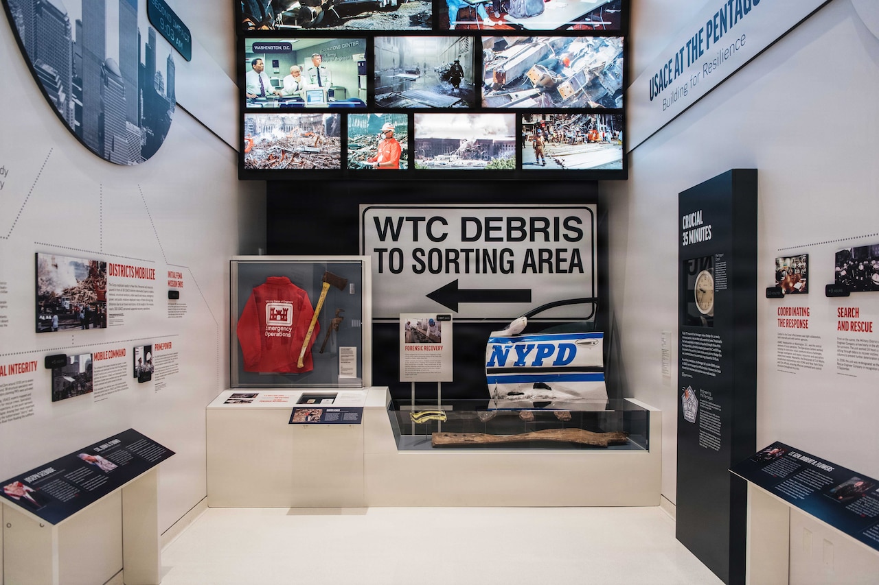 A damaged door from an NYPD squad car, a street sign that says "WTC Debris To Sorting Area" and TV screens showing images from 9/11 hang on a wall in a museum.