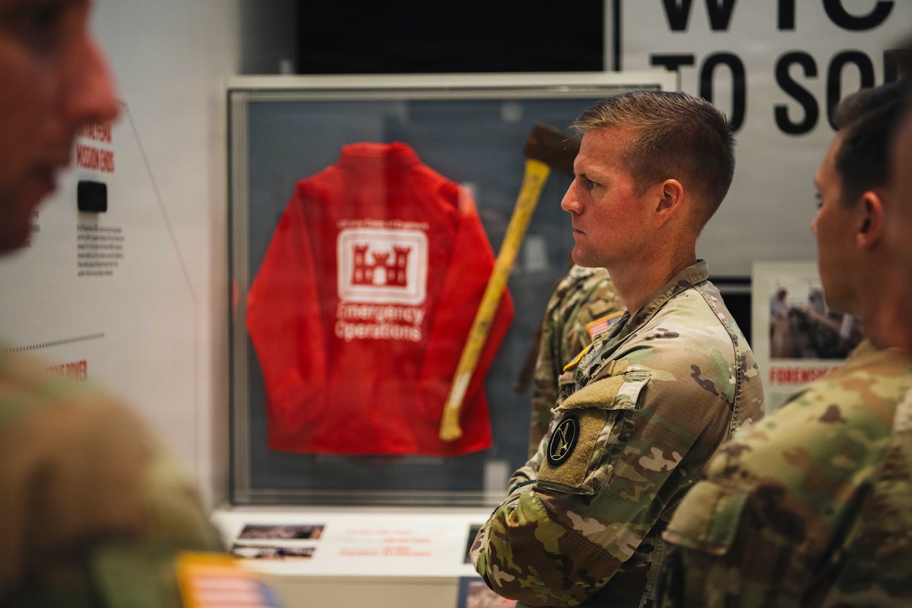 People in camouflage uniforms look at artifacts and timelines on the walls of a museum exhibit. A red jacket and an ax are in a glass case in the background.