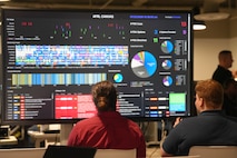 two men sit in chairs talk about the large computer display in front of them