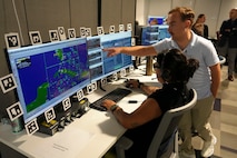 a woman wearing a headset, specialty glasses and watch works at a computer while a man stands beside her pointing out something on the screen