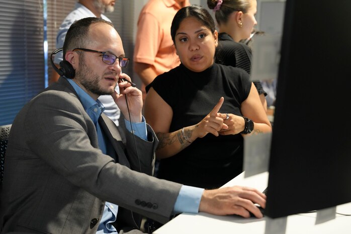 a man wearing a headset works on a computer while a woman stands and explains info on the screen.