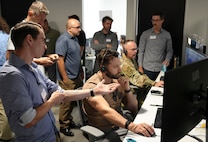 a group of people stand behind observing a US military member and civilian working on computers