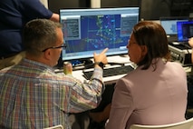 a woman works on a computer while a man sits beside her explaining the info on the screen.