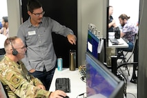 a US military member wearing a headset works on a computer while a man stands beside him pointing out something on the screen