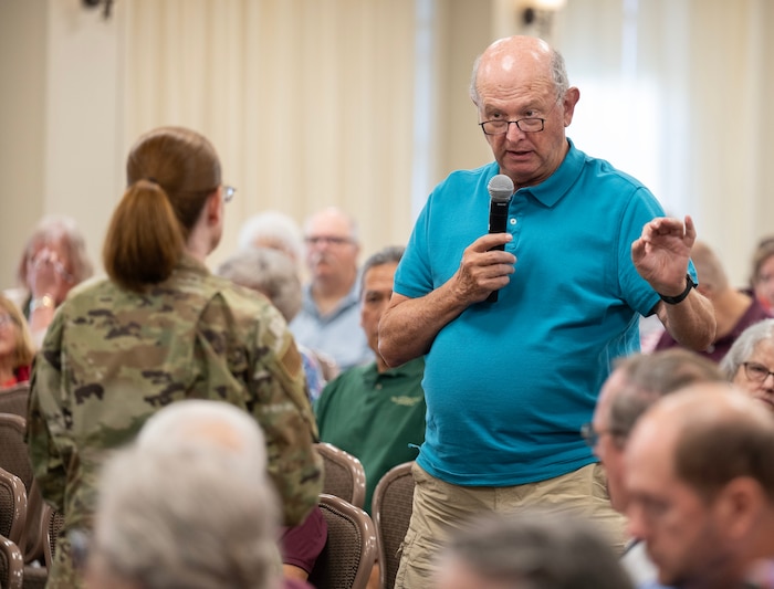 An elderly man holds a microphone with one hand and gestures with the other as he talks to a woman in camouflage uniform .