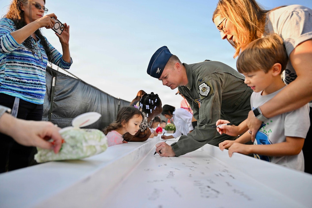 An airman in a green jumpsuit leans over and uses a black marker to sign a white slab outside as a woman uses a phone to take a photo and other women and children look at signatures on the slab.