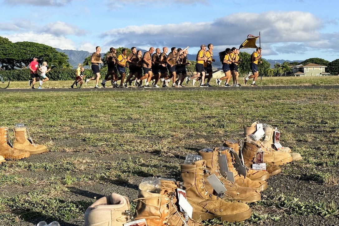 A pack of people in athletic attire and a few children run behind a person holding a flag on a pathway next to a field with rows of work boots that have been tagged with photos of people.
