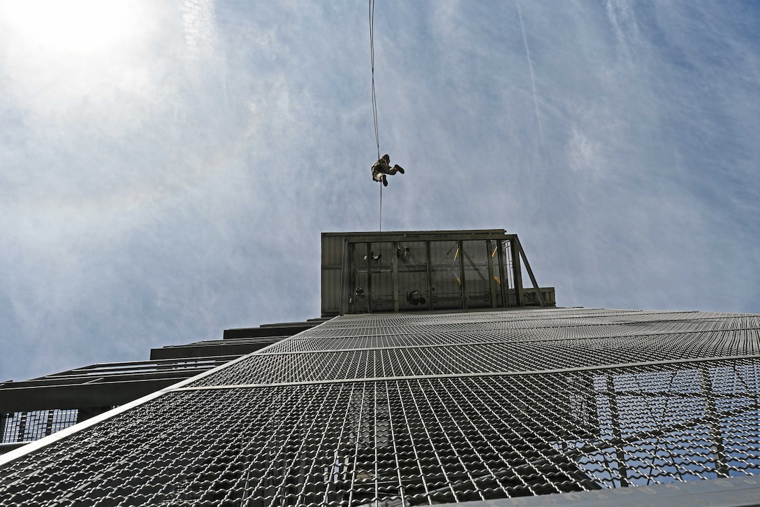 An upward view of a person using a long rope to rappel from a tall metal platform during daytime, with a blue sky above.