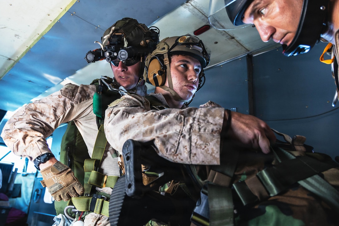 A Marine wearing parachute gear, a helmet and eye protection stands behind a Marine wearing a helmet and ear protection and holding a large duffel-like bag and rifles, as another person wearing a helmet looks down.