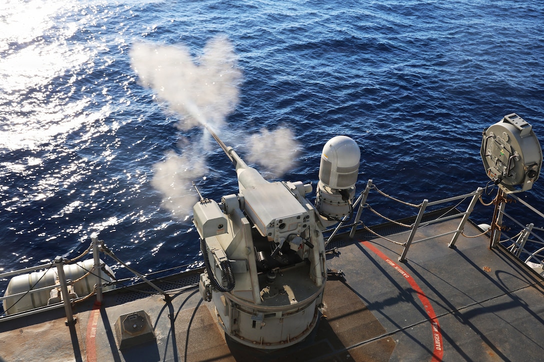 Smoke wafts as a large gun is fired on the deck of a ship at sea during daytime.