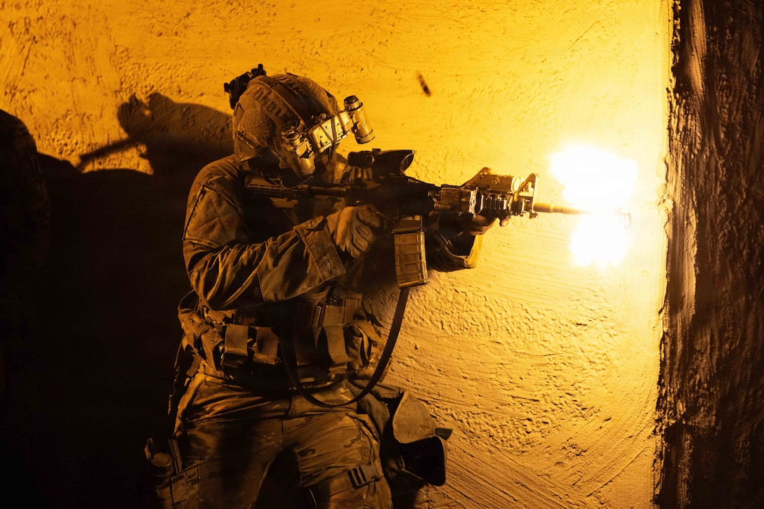 A soldier in combat gear fires a rifle while standing against a wall in dark conditions, creating an orange blast from the gun nozzle.