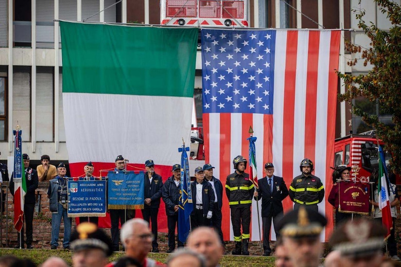 A dozen men wearing business attire and firefighter uniforms stand on a stage holding flags and signs; there are two large American and Italian flags in the background.