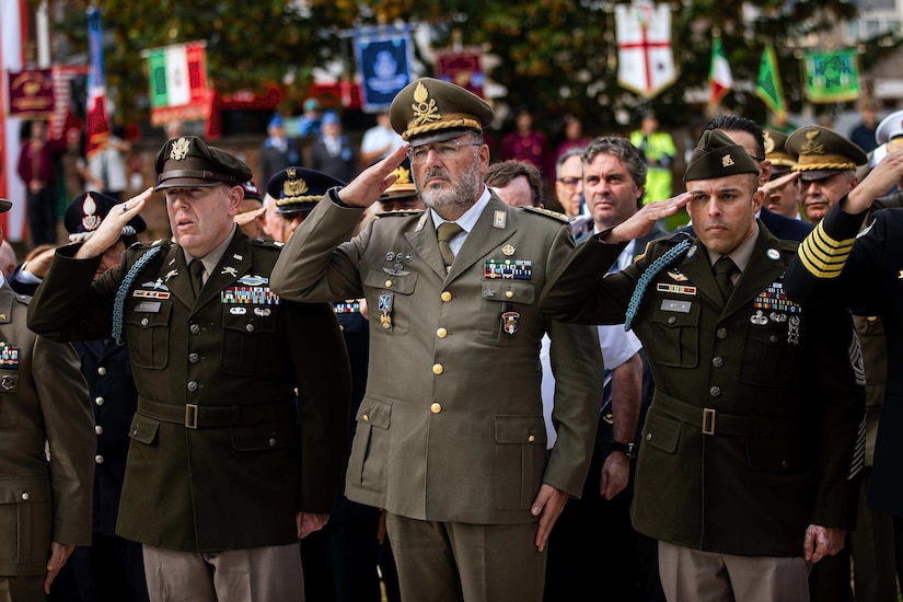 A dozen men wearing military dress uniforms stand and salute outside.