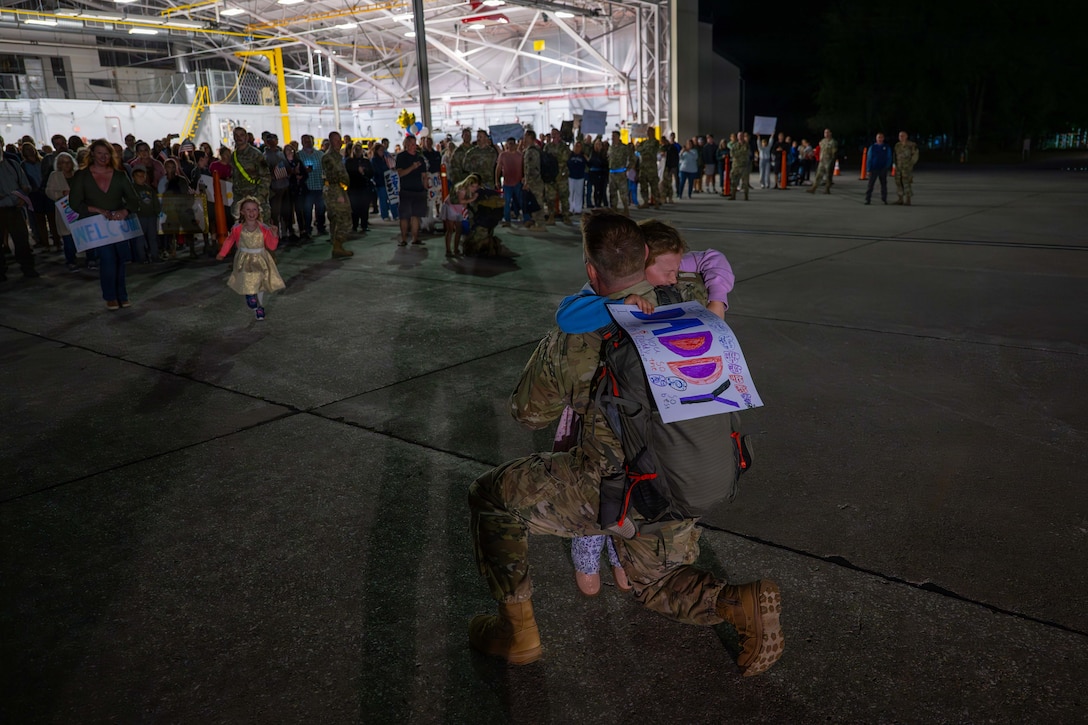 A father in a camouflage uniform  kneels to hug a child who is holding a sign that says "Daddy" in bright colors outside in front of an aircraft hangar, as another child runs toward them.