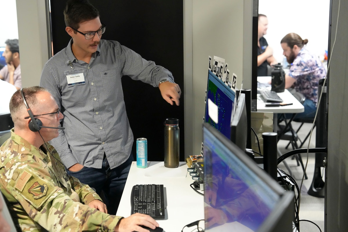 a US military member wearing a headset works on a computer while a man stands beside him pointing out something on the screen