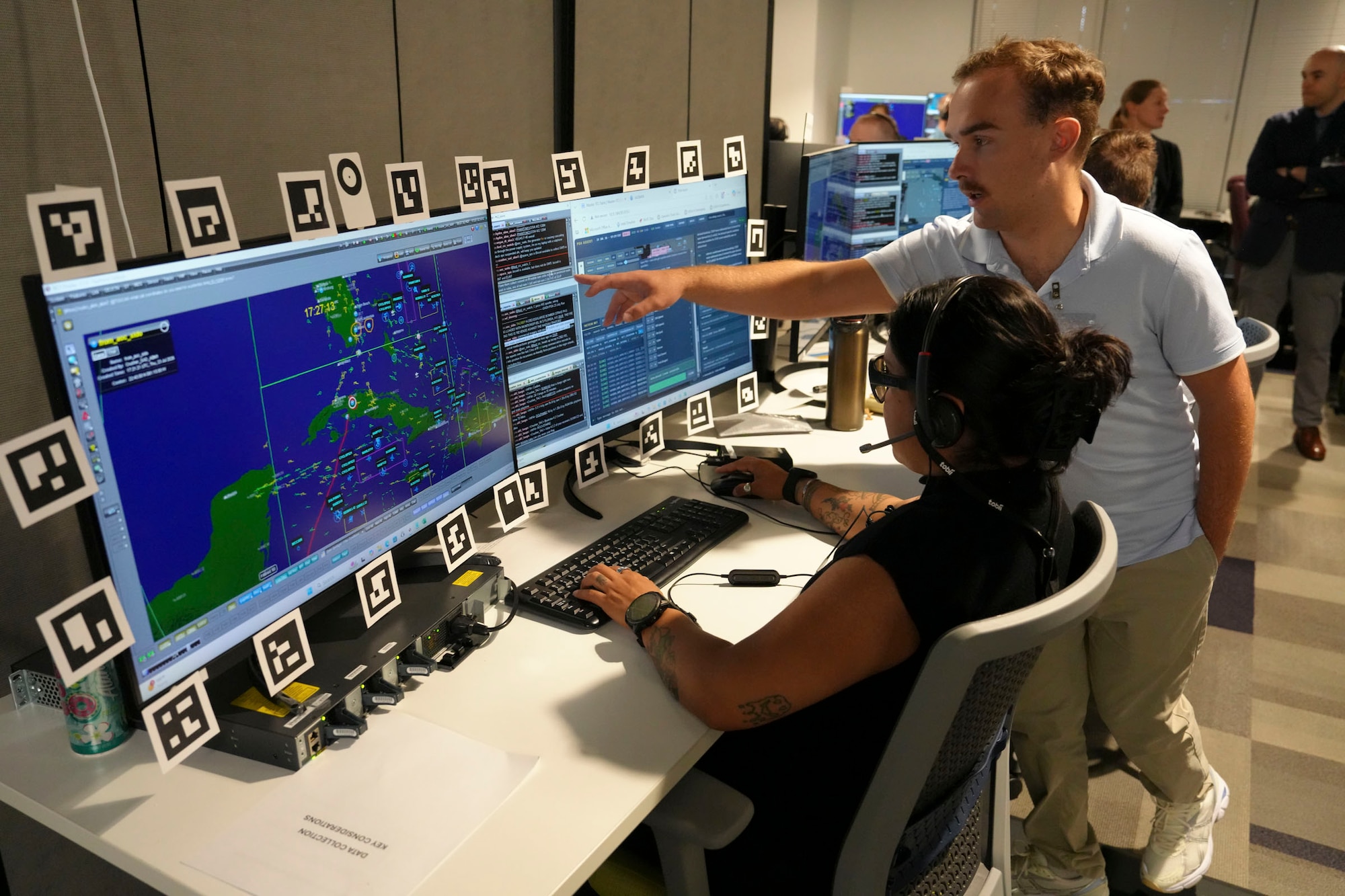 a woman wearing a headset, specialty glasses and watch works at a computer while a man stands beside her pointing out something on the screen