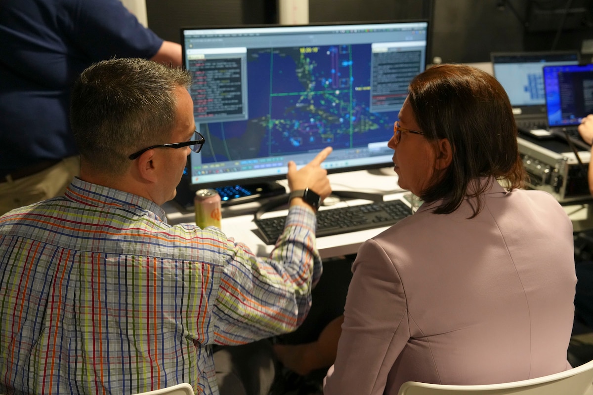 a woman works on a computer while a man sits beside her explaining the info on the screen.